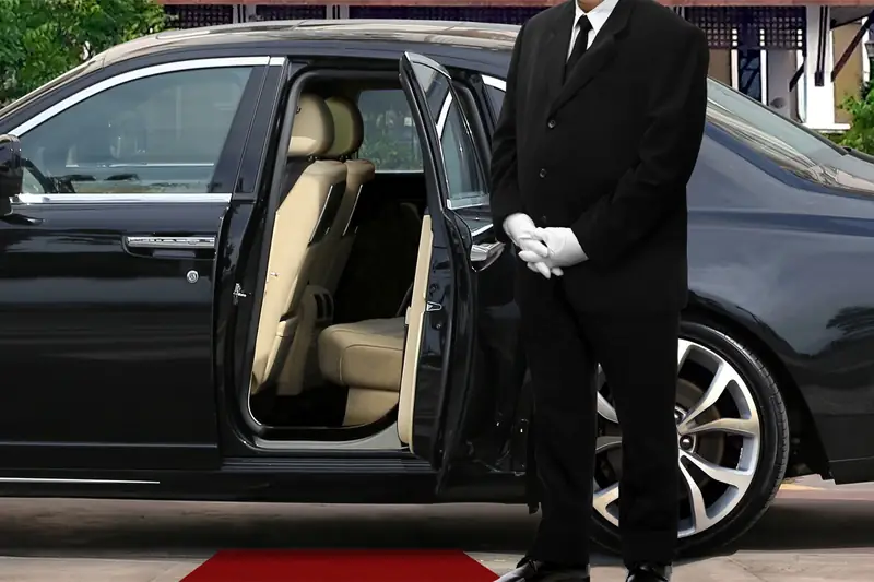 A chauffeur in white gloves beside the open door of the car he drives.